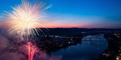 Fireworks burst over a river at dusk, city lights in the distance.