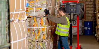 Worker in a high-vis vest moves stacked, bound cardboard in a warehouse.
