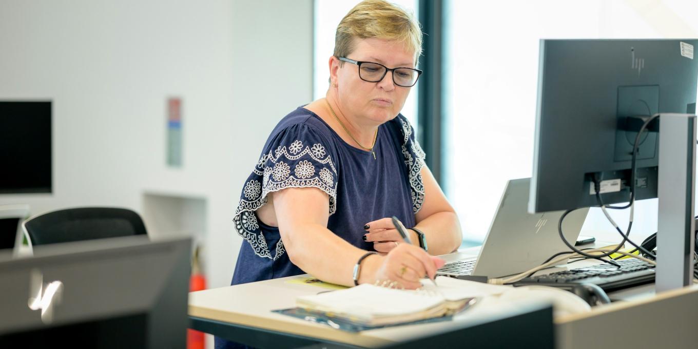 Woman working at a desk with a computer and paperwork, focused and wearing glasses.