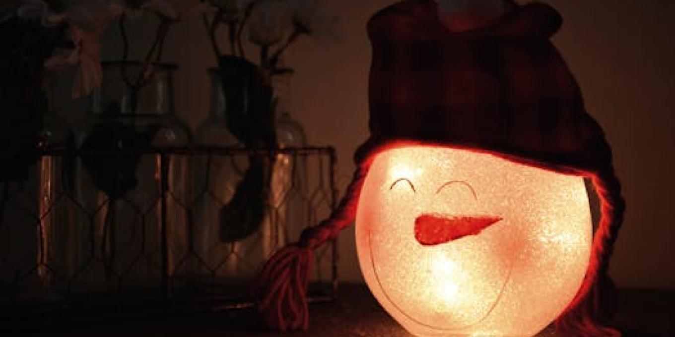 Glowing snowman lamp with a warm hat, surrounded by festive jars indoors, creating a cozy winter atmosphere.