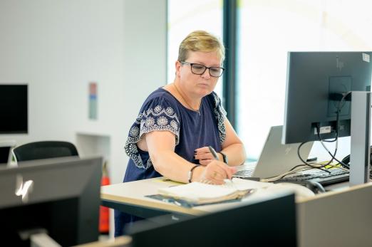Woman working at a desk with a computer and paperwork, focused and wearing glasses.