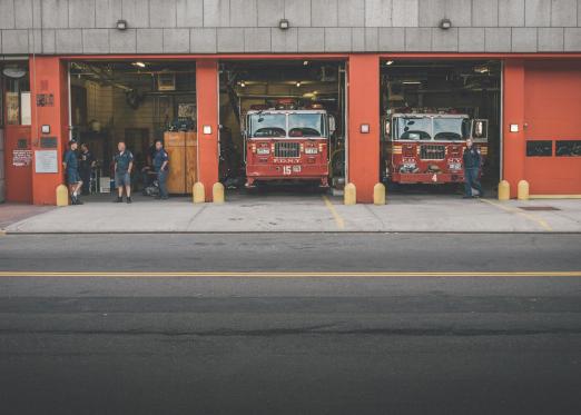 Fire trucks parked in a station with firefighters standing nearby.