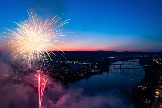 Fireworks burst over a river at dusk, city lights in the distance.