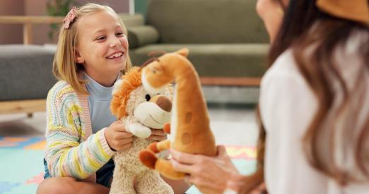 Child smiling, holding a plush lion, facing a woman with a plush giraffe.