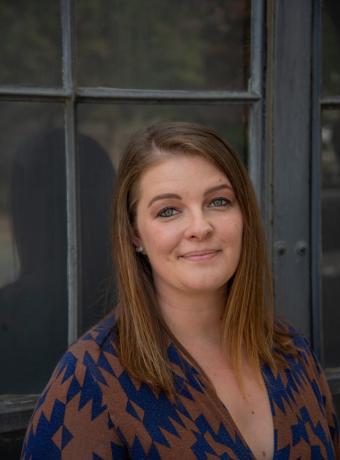 Smiling woman in a patterned shirt, standing by a window.